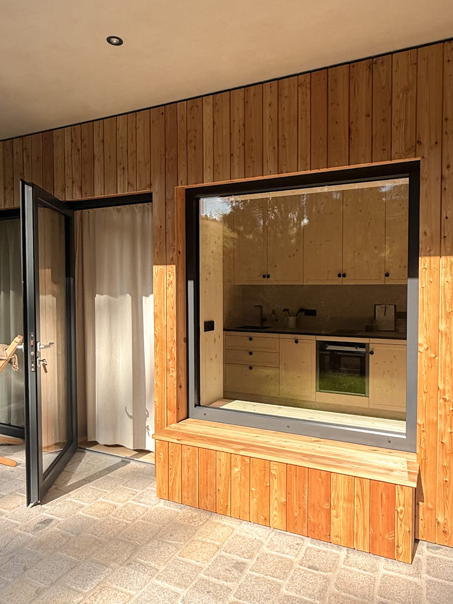 Das Bild zeigt ein modernes Tiny House in natürlicher Umgebung. Die Fassade besteht aus hellen, vertikal angeordneten Holzbrettern, und das Dach ist leicht geneigt mit einem dunklen Randabschluss. Vor dem Haus befinden sich zwei gemütliche Liegestühle, die mit Fellen belegt sind, und daneben steht ein kleiner Holzhocker. Die Stimmung ist ruhig und einladend – ideal für Entspannung in der Natur.