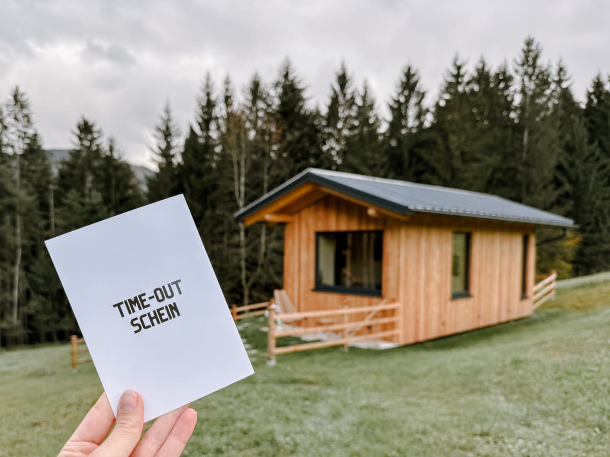 ine Person hält einen Gutschein mit der Aufschrift "TIME-OUT SCHEIN" vor einem modernen, aus Holz gebauten Tiny House, umgeben von grünen Wiesen und Bäumen.