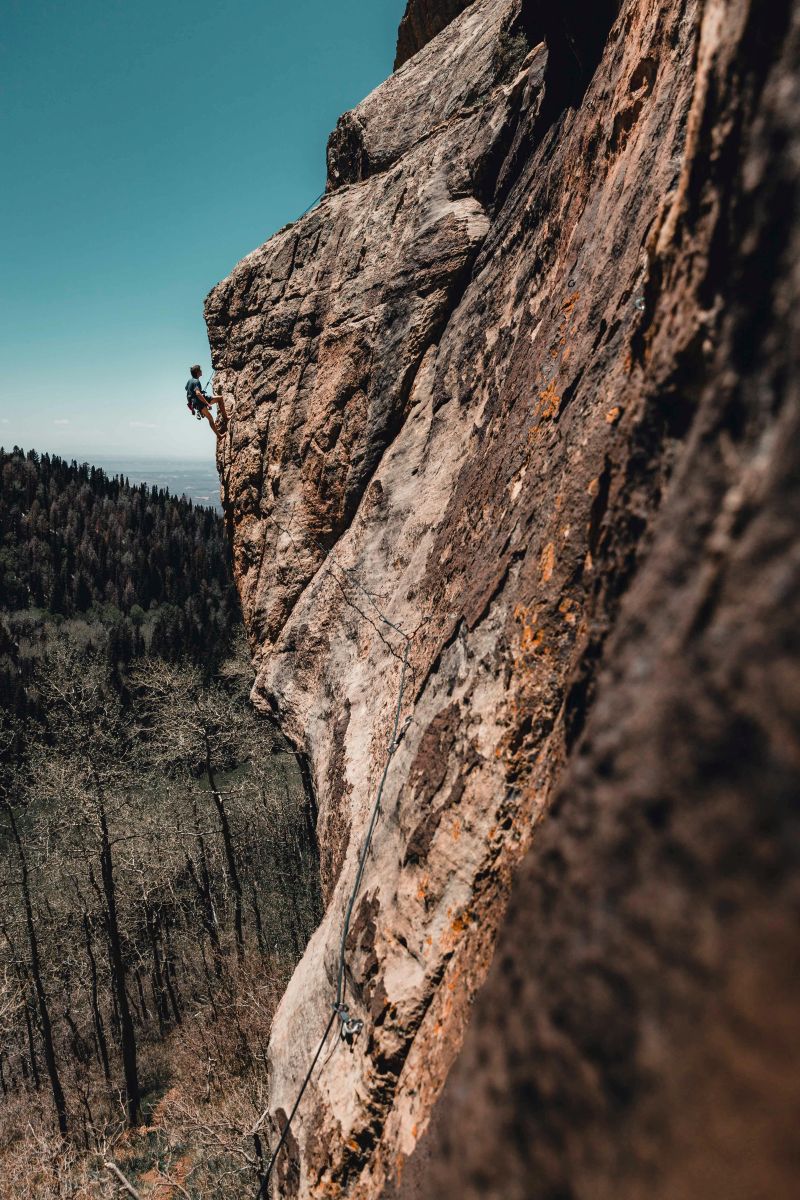 Ein Kletterer schwingt sich an einem steilen Felsen entlang, während er eine schwierige Kletterroute bewältigt. Der Himmel ist klar und der Wald ist weit im Hintergrund zu sehen.
