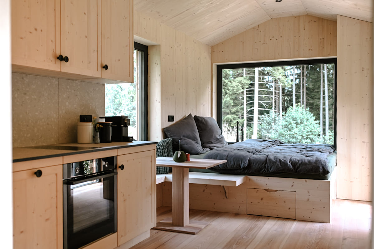 nnenansicht eines Tiny Houses mit einer modernen Küche und einem Bett, das von einem großen Fenster mit Waldblick umgeben ist.