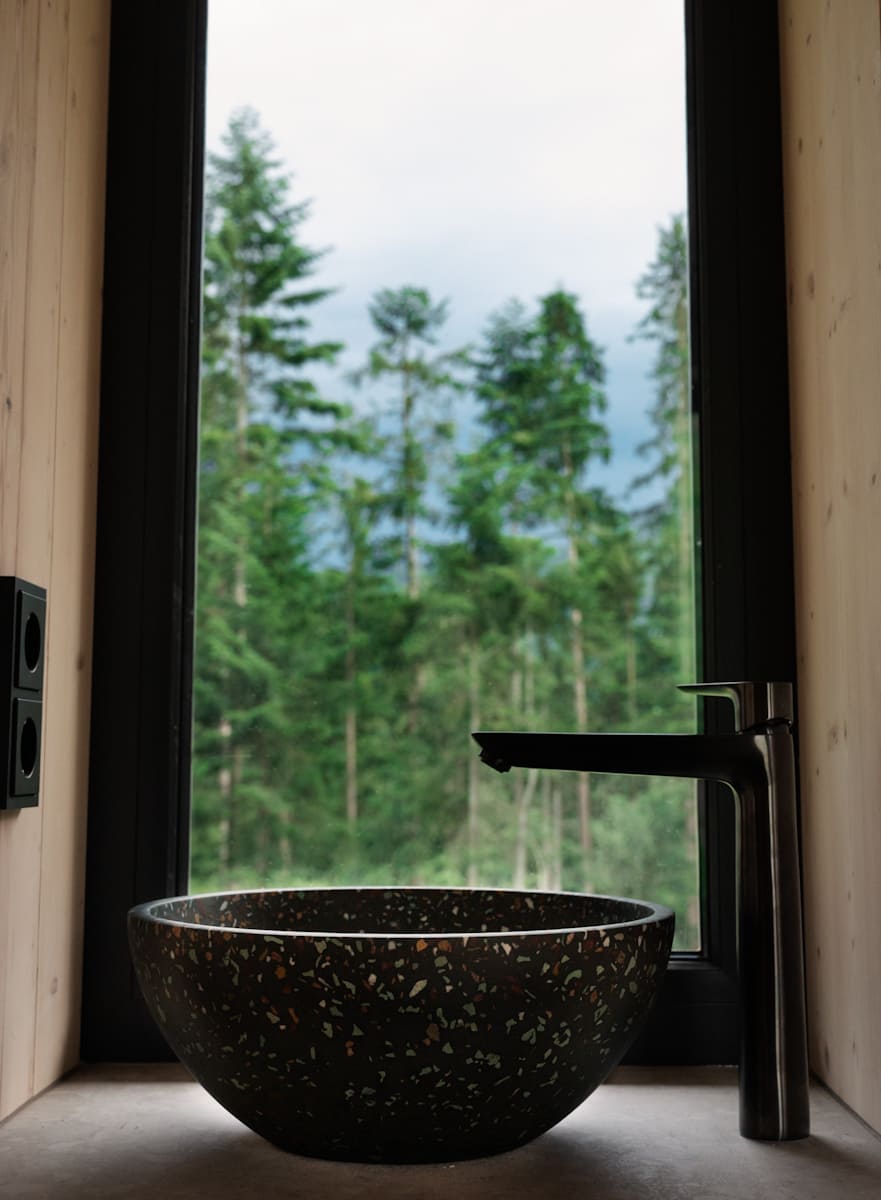 in modernes Badezimmer mit einem schwarzen Waschbecken aus Terrazzo und einem stilvollen Wasserhahn. Durch das große Fenster sieht man den grünen Wald im Hintergrund.
