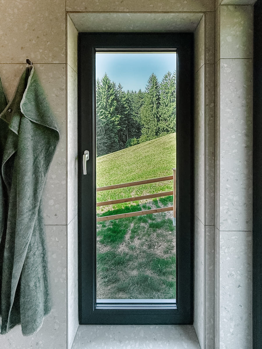 Ein gemütliches Schlafzimmer im Tiny House mit einem Bett, weiße Bettbezüge, Kissen und einer Decke, und einem großen Fenster, das den Blick auf den grünen Wald im Mostviertel freigibt.